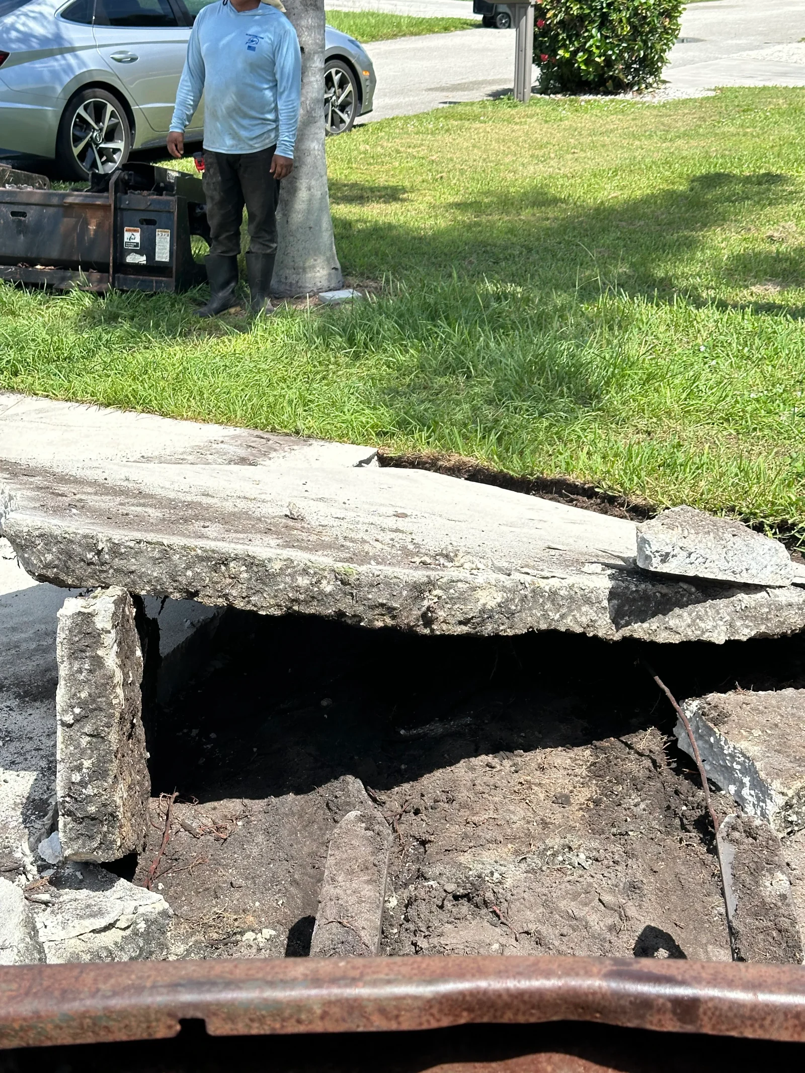 Broken concrete sidewalk slabs removed to expose excavated soil trench with rusted steel plate and skid steer loader