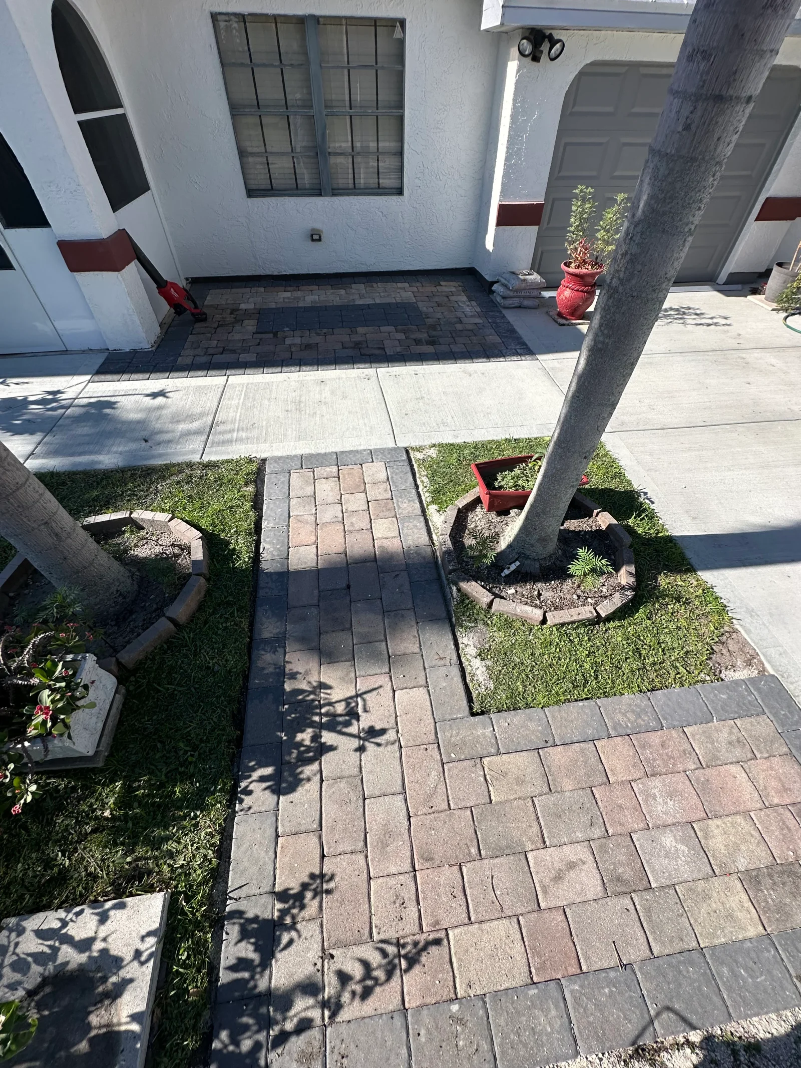 Multi-tone interlocking paver walkway with dark border and framed paver landing beside a concrete driveway and palm tree planters