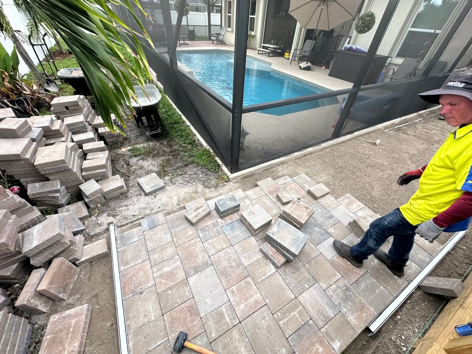 Interlocking concrete pavers being laid on sand base with aluminum edge restraint beside screened pool enclosure