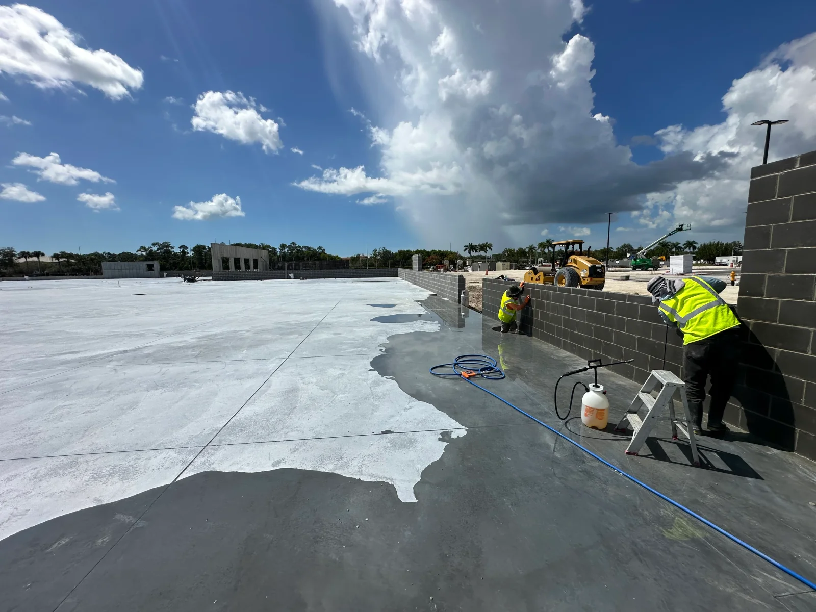 Crew sealing a large concrete slab and applying coating to a gray concrete block wall with a pump sprayer and blue hose