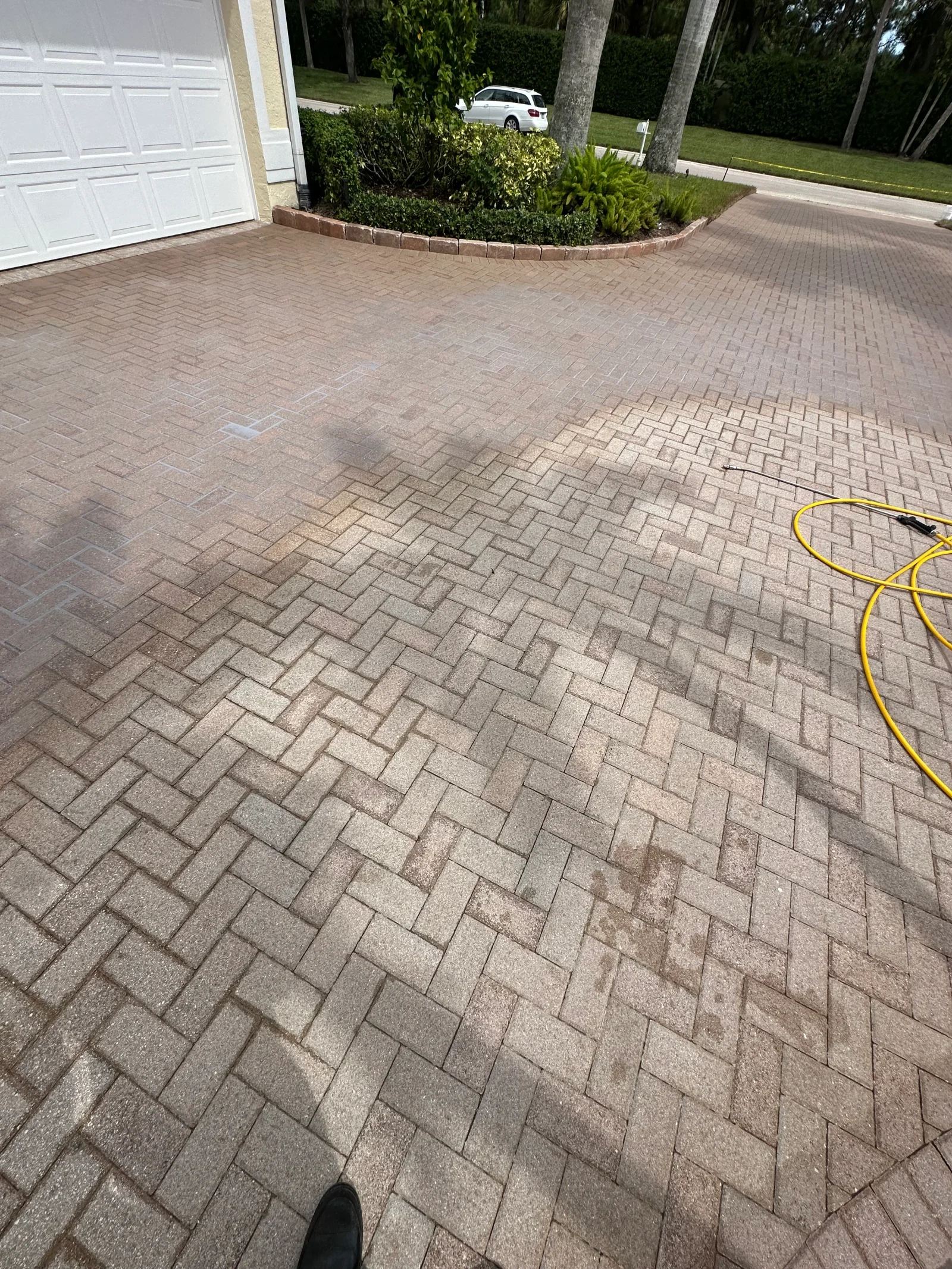 Herringbone brick paver driveway partially pressure washed, with yellow hose and wand on the right and clean versus stained pavers