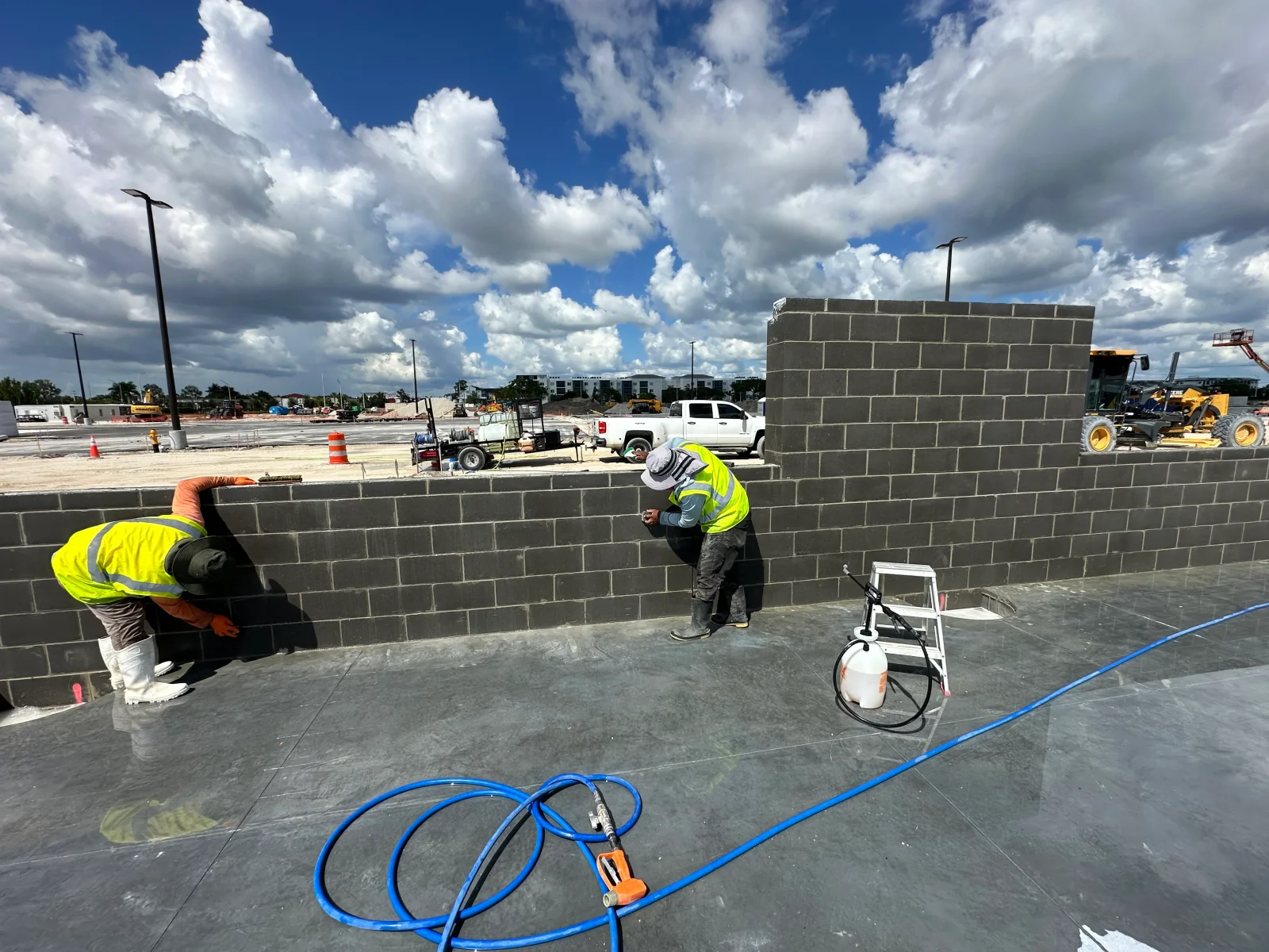 Workers laying dark gray concrete block wall on smooth concrete slab with blue hose and sprayer tank nearby