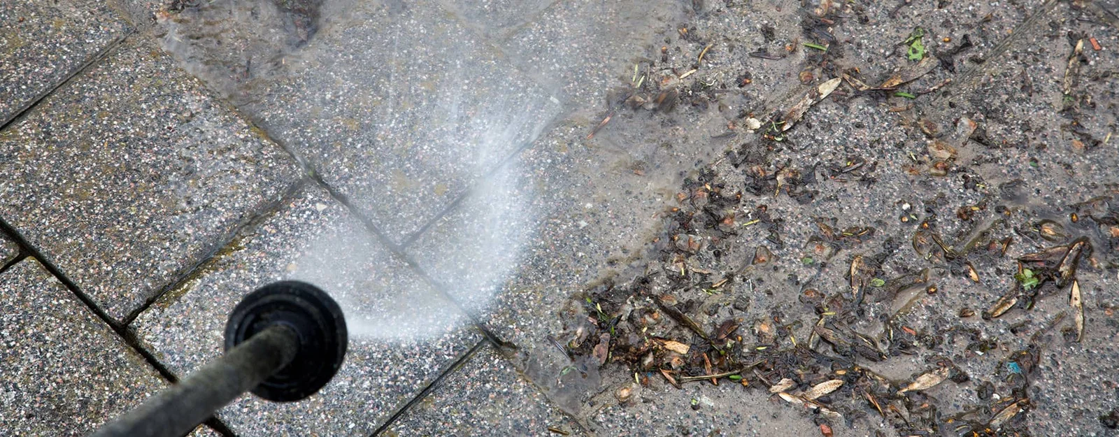 Pressure washer wand cleaning gray textured concrete pavers, with debris and leaves remaining on the uncleaned side