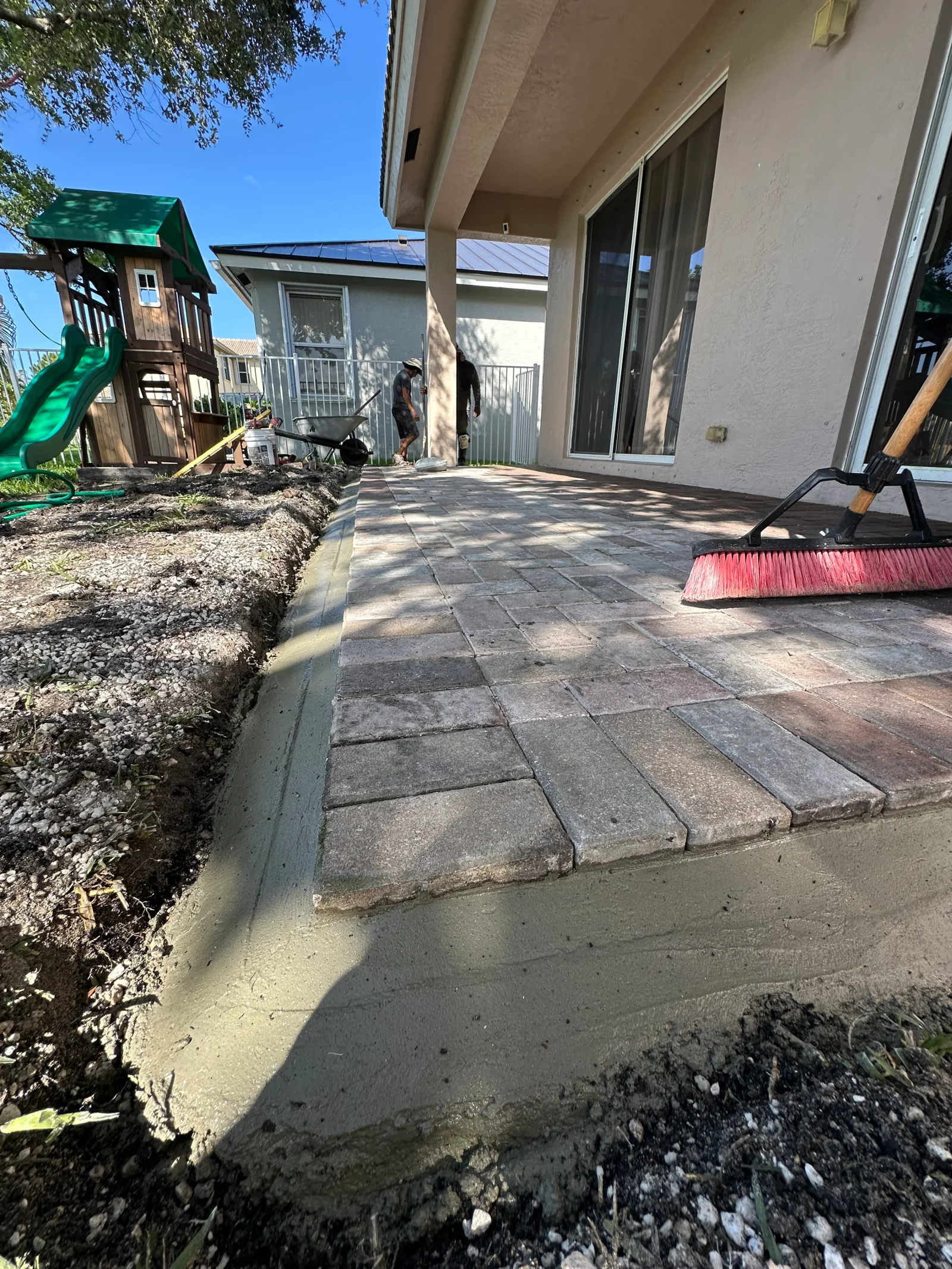 Multi-toned rectangular concrete pavers being laid with a fresh concrete edge restraint along a stucco home's patio