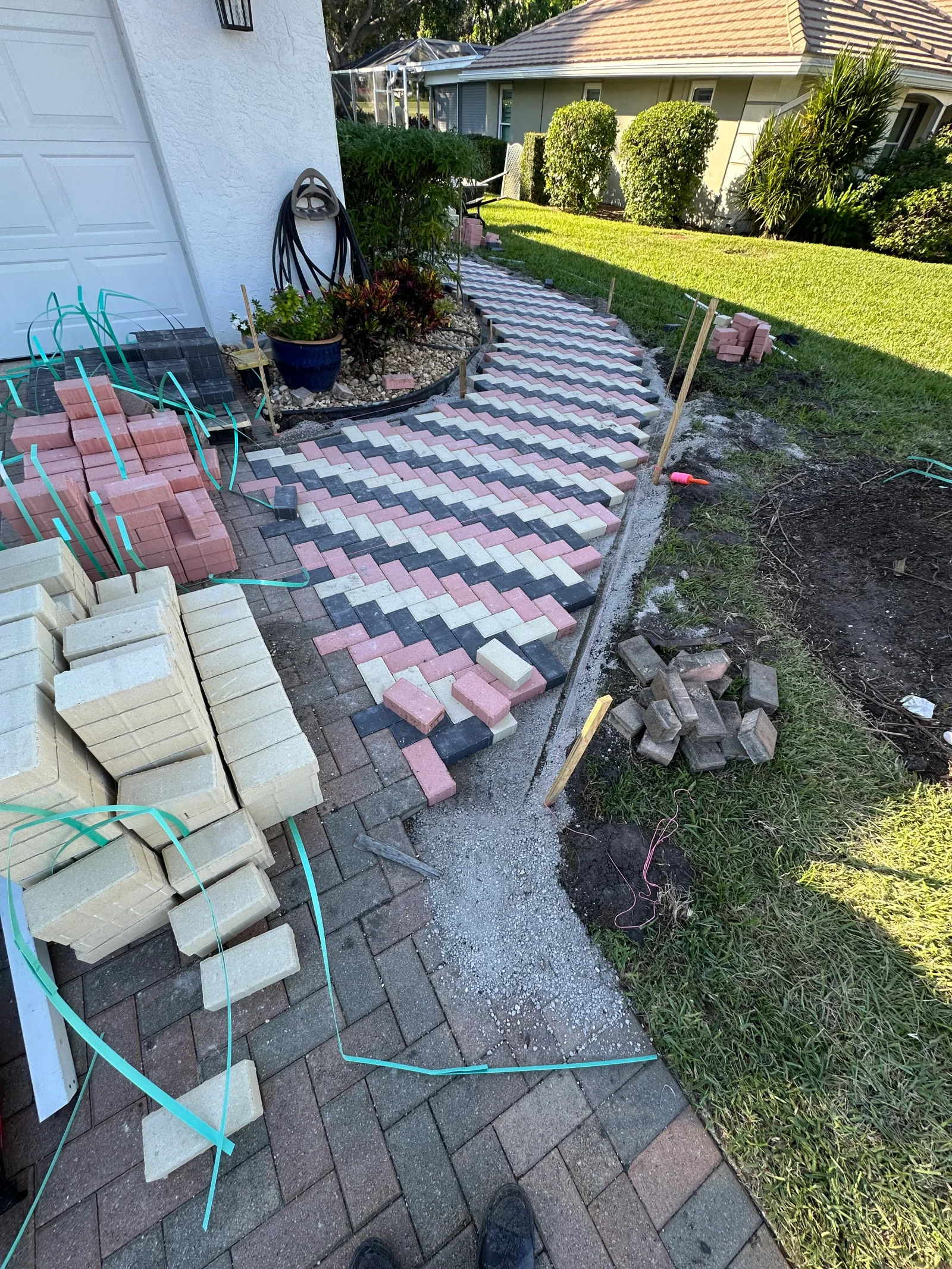 Interlocking pink, gray, and cream concrete pavers laid in a herringbone pattern on a curved walkway with wooden stakes