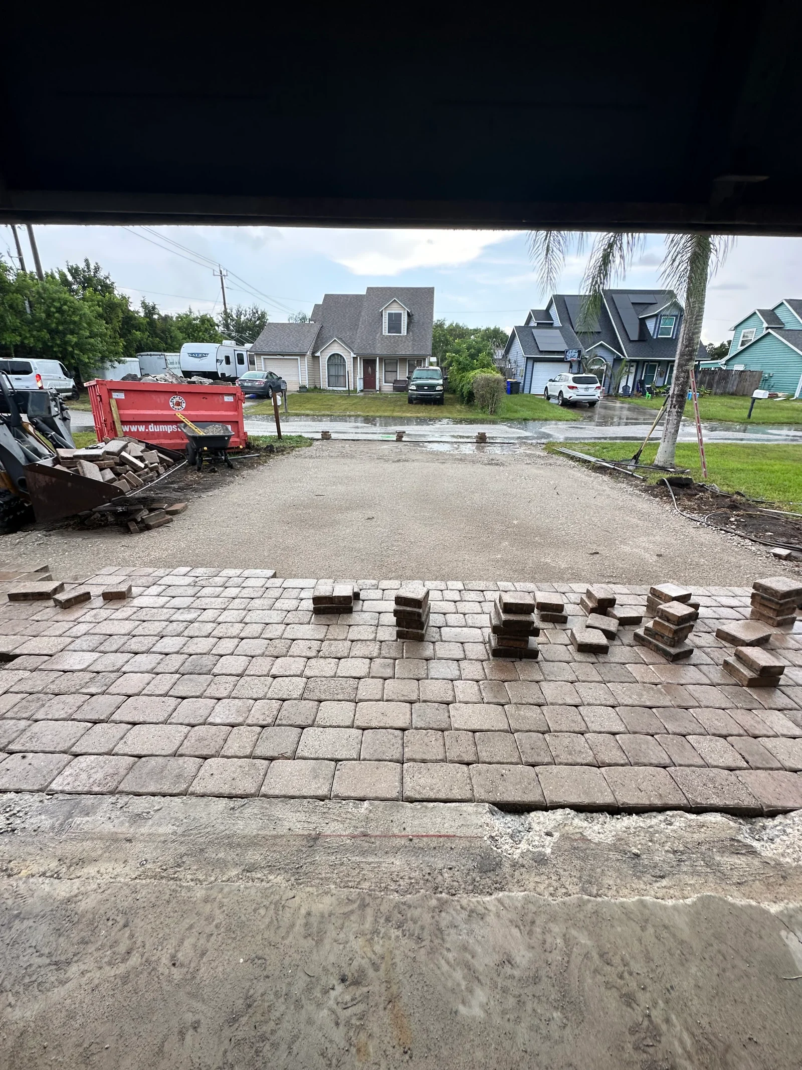 Interlocking concrete pavers in brown and tan laid over a gravel base with stacked pavers and skid steer on site