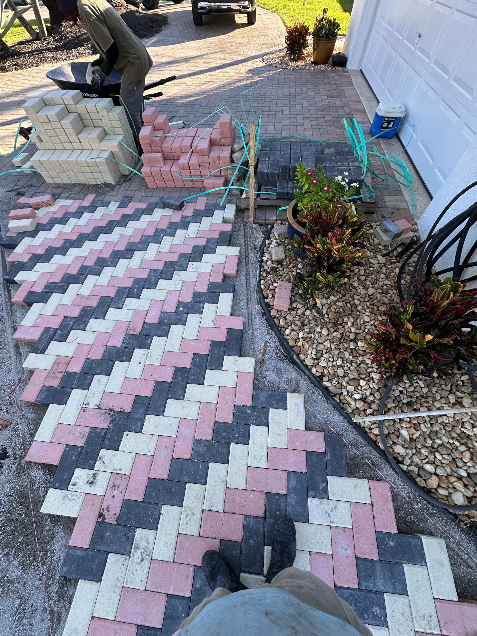 Crew laying cream, pink, and charcoal herringbone concrete pavers with string lines beside a curved black edging and river rock bed